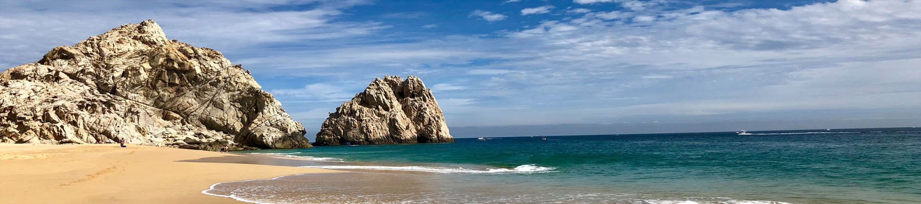 Picture of a beautiful beach in Cabo San Lucas, Mexico.  The picture shows tall rocks, a sandy beach, blue water and a blue sky with white clouds