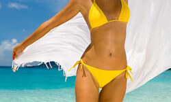 Picture of a woman, happy with her abdomen and waist  liposuction procedure she had with Top Plastic Surgeons in beautiful Cabo San Lucas, Mexico.  The woman is wearing a two piece yellow bathing suit and walking through the surf at the beach.