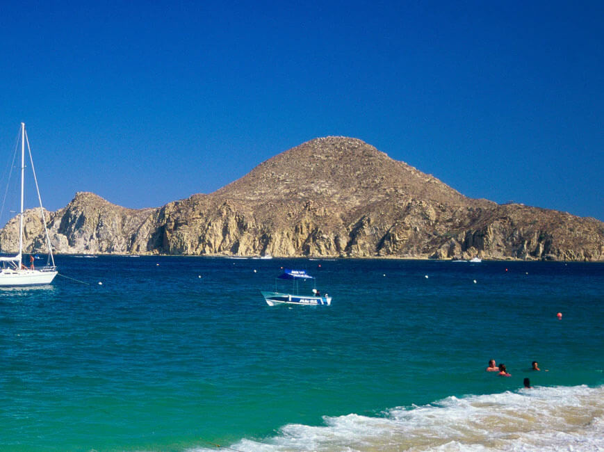 Picture of a beautiful sandy beach in Cabo San Lucas, Mexico.  The picture shows white sand, blue water and framed by tall rocks behind the shoreline.