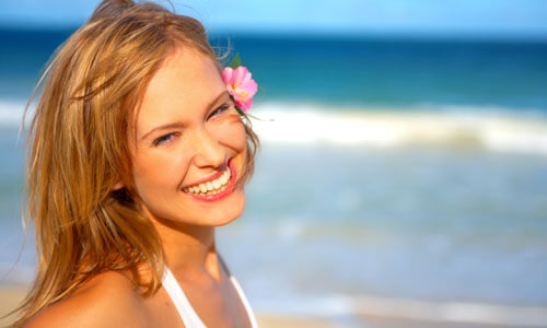 Close-up picture of a beautiful woman, happy with her face lift with neck lift in Cabo San Lucas, Mexico.  The woman has long sandy blonde hair and is looking directly at the camera with the ocean behind her.