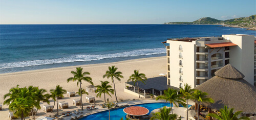 Picture of a beautiful Los Cabos beach with blue water and a hotel and swimming pool featured in the foreground.  The pool is oval shaped with palm trees all around it