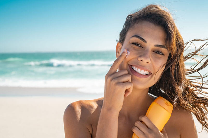 Picture of a smiling woman, happy with her nose surgery she had at Cabo MedVentures.  The woman has long brown hair and is standing on a sandy Cabo beach with the ocean in the background.