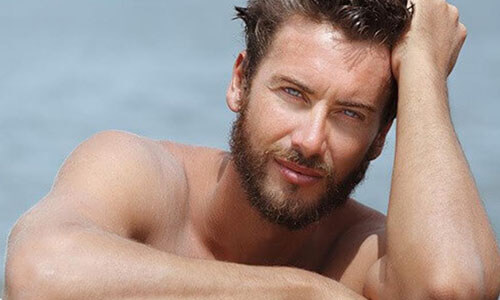 Close-up picture of a man,  happy with his hair transplant  procedure he had in Cabo San Lucas, Mexico.  The man is shown leaning against the edge of a pool and looking directly at the camera with an arm raised to his head to feature his hair transplant.