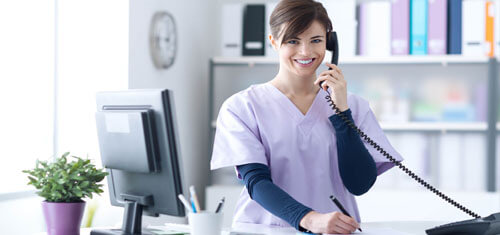 Picture of a receptionist representing Top Plastic Surgeons in beautiful Cabo San Lucas, Mexico.  The woman has short brown hair, is wearing a hospital smock and is standing at the receptionist desk while smiling at the camera.